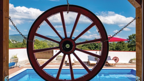 A large wooden wheel with chains, overlooking a swimming pool and outdoor seating area with a view of mountains. - Stunning home in Gospic with sauna (Gospic)