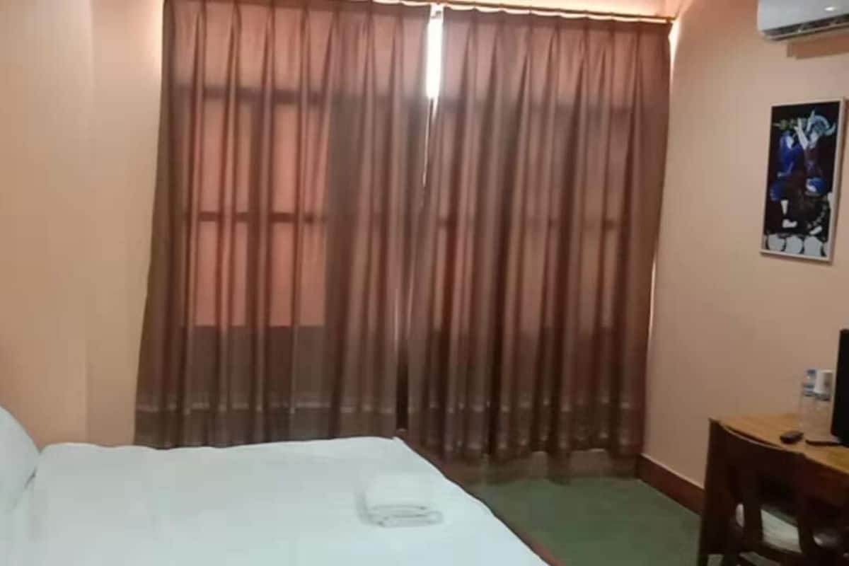 A hotel room with a bed, a wooden headboard, a window with sheer curtains, a small table with a chair, and a poster on the wall.
