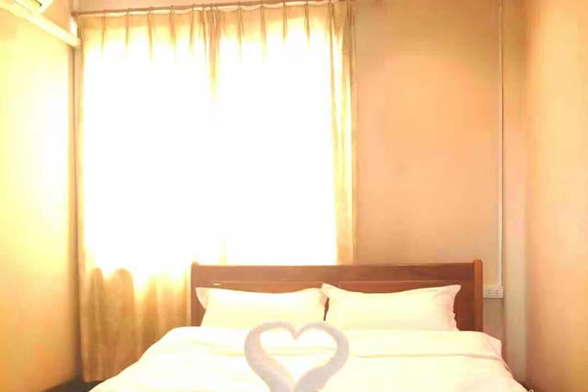 A neatly made bed with a heart-shaped towel arrangement, a wooden headboard, and a window with curtains.