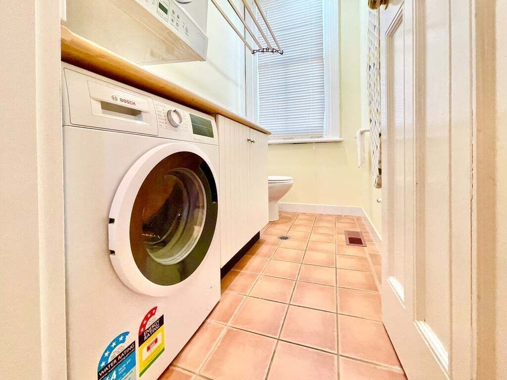 A compact laundry area with a Bosch washing machine, a toilet, and a window with blinds.