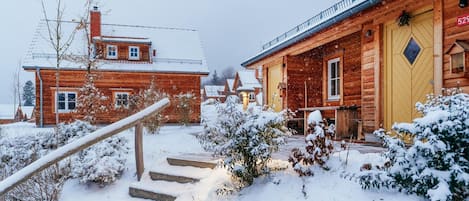 A wooden cabin with a yellow door, a staircase leading to the entrance, and snow-covered surroundings.