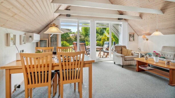 En rummelig loftrum med højt til loftet, træbjælker, et spiseområde med træstole, en sofa og et sofabord. Der er en skydedør af glas, som fører til en udendørs have.
