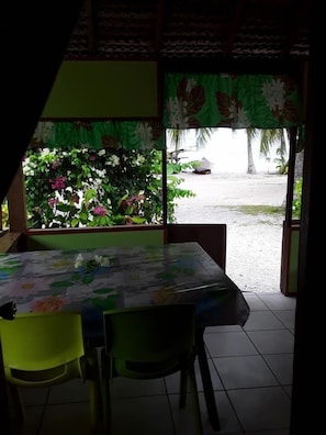 A room with a table covered by a floral tablecloth, two chairs, and a view of a beach through a window with a green curtain. - Pension Chez Glorine (Rangiroa)