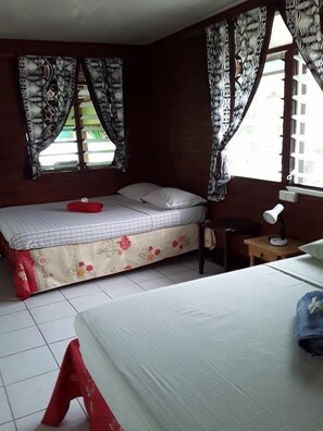 A room with two beds, one with a floral bedspread and the other with a plain white bedspread. There is a small table with a lamp and a chair. - Pension Chez Glorine (Rangiroa)
