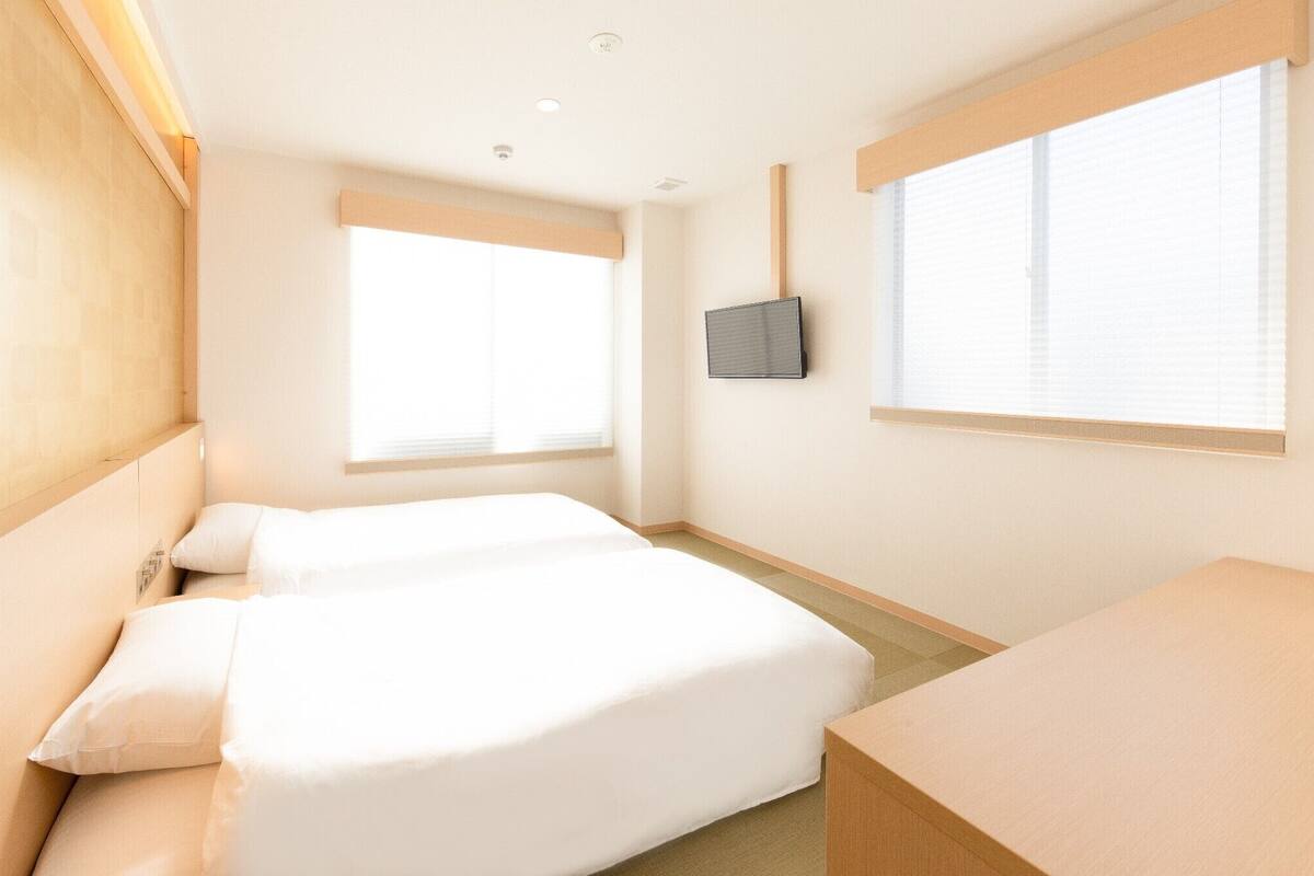 A hotel room with two beds, a TV mounted on the wall, and a window with blinds.