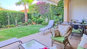 A patio with wicker furniture, a glass table, and a lush garden with a variety of plants and a palm tree. - Mijas Costa Lovely Holiday Home for 6 (Mijas Costa)