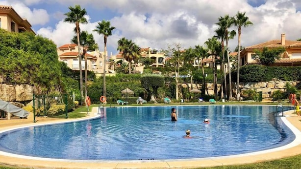 A swimming pool surrounded by palm trees and a stone wall, with a clear sky above. - Mijas Costa Lovely Holiday Home for 6 (Mijas Costa)