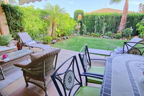 An outdoor patio with wicker furniture, a tiled table, and a well-maintained garden with various plants and a lawn.