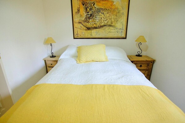 A bed with a white and yellow bedspread, two bedside lamps, a painting of a leopard on the wall, and a wooden nightstand with a drawer.