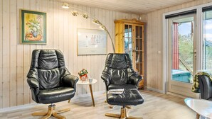 A room with two leather armchairs, a small table with a vase, a floor lamp, a wooden cabinet, and a large window with a view of the outdoors.