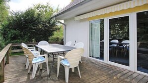 Um deck de madeira com mesa e cadeiras, uma porta de vidro deslizante e uma parede externa branca.