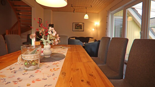 Uma sala de jantar com uma mesa de madeira, cadeiras, um vaso com flores e uma escada ao fundo.