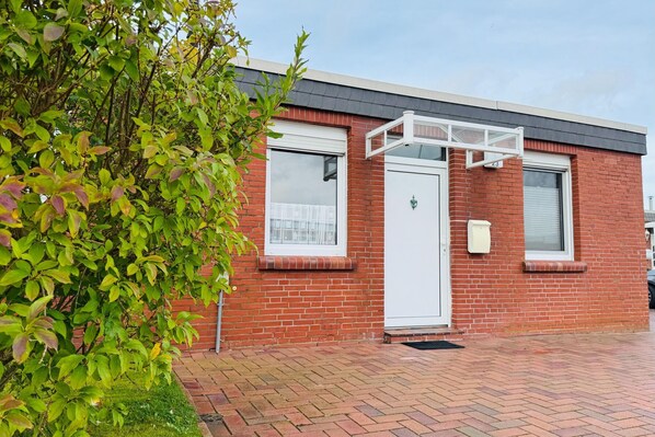A red brick building with a white door, windows, and a small porch. - Bungalow, Dornumersiel (Dornumersiel)