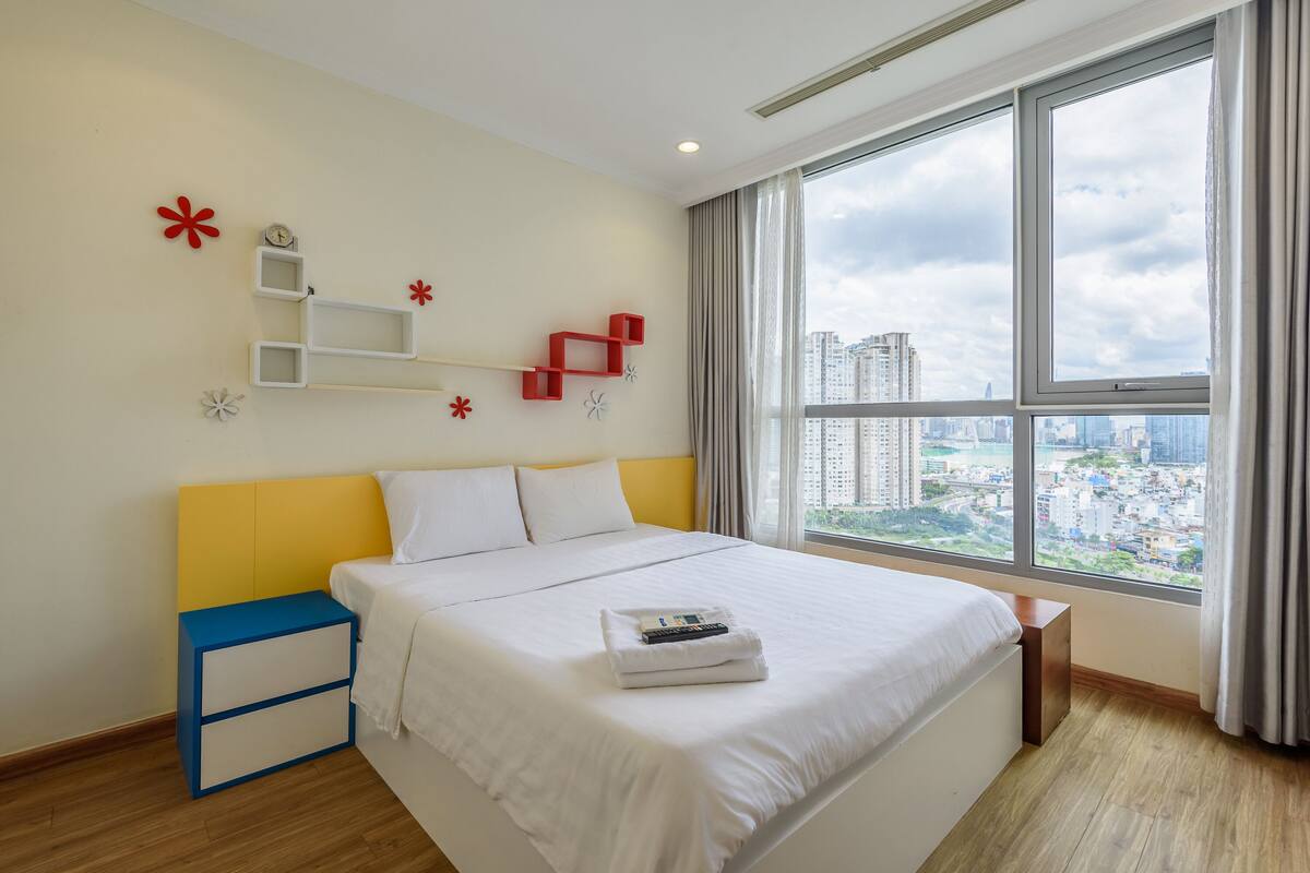 A modern hotel room with a large bed, a bedside table, a nightstand, and a view of the city through large windows.