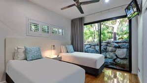 A bedroom with two beds, a ceiling fan, a TV, and a view of a stone wall and greenery. - Pinnacle 4, Seaview Apartment on Hamilton Island (Hamilton Island)