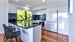 A modern kitchen with a central island, white cabinetry, and a view of greenery through large windows. - Pinnacle 4, Seaview Apartment on Hamilton Island (Hamilton Island)