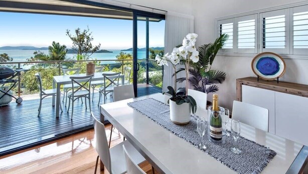 A modern dining area with a view of the sea, a balcony with outdoor furniture, and a table set with a bottle and glasses. - Pinnacle 4, Seaview Apartment on Hamilton Island (Hamilton Island)
