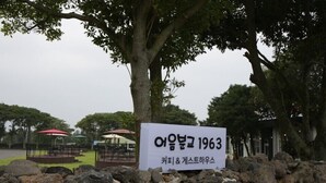A stone wall with a sign in Korean, a tree, and outdoor seating areas in the background. - Jeju Eoeumbungyo 1963 Guesthouse (Jeju City)