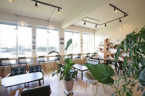A spacious cafe with large windows, wooden tables, and chairs, and a variety of plants. - Jeju Eoeumbungyo 1963 Guesthouse (Jeju City)