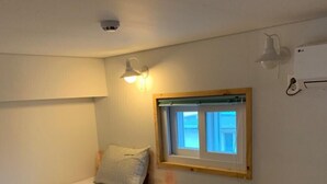 A small, sparsely furnished room with a single bed, a window, and a ceiling light. - Jeju Eoeumbungyo 1963 Guesthouse (Jeju City)