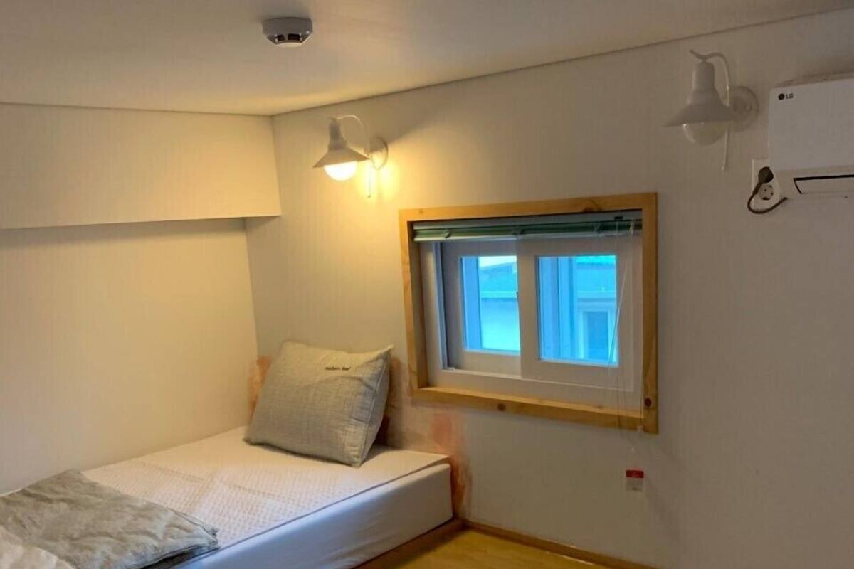 Et lille, nødtørftigt møbleret værelse med en enkelt seng, et vindue og en loftlampe.