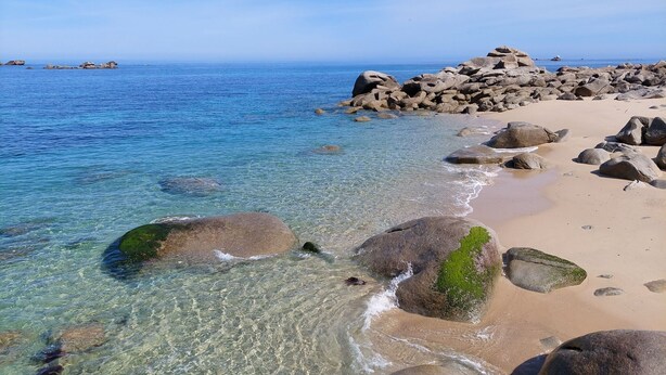 A beach with clear water, large rocks, and a distant view of the sea.