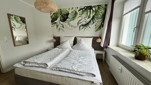 A neatly made bed with a quilt, a large mirror, a tropical leaf mural, bedside lamps, and a window with a plant.