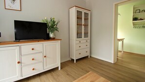 A room with a flat-screen TV on a wooden cabinet, a white cabinet with drawers, a glass-fronted cabinet, and a small table with a plant.