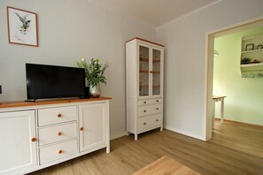 Une pièce comprenant un téléviseur à écran plat posé sur un meuble en bois, un meuble blanc avec des tiroirs, un meuble avec des portes en verre et une petite table sur laquelle se trouve une plante.