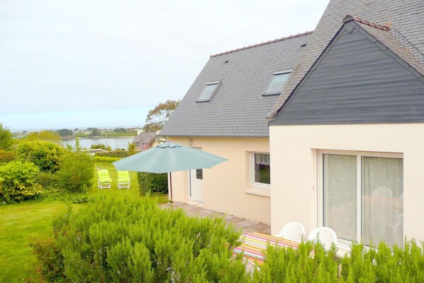 Ein Haus mit geneigtem Dach, einer Terrasse mit Stühlen und Sonnenschirm, und ein Blick auf ein Gewässer im Hintergrund.