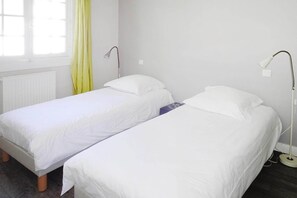 Deux lits simples avec du linge de lit blanc, une fenêtre avec des rideaux jaunes et un radiateur au mur.