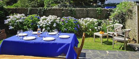 Un área de comedor al aire libre con una mesa dispuesta para cuatro personas, rodeada de abundante follaje y plantas en flor.