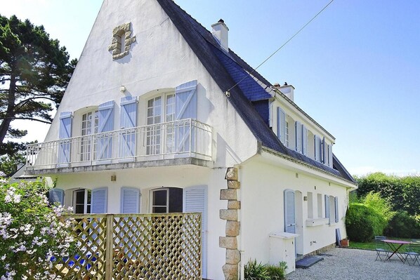 Un bâtiment blanc doté d’un toit à forte pente, d’un balcon avec des balustrades blanches et d’un escalier en pierre menant à l’entrée.