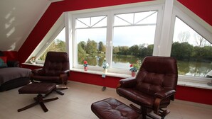 Une pièce avec un plafond en pente, une grande fenêtre donnant sur un lac, et deux fauteuils en cuir assortis de deux poufs.