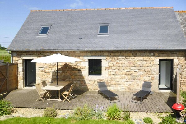 Une maison en pierre avec une terrasse en bois, une table et des chaises, ainsi que deux fauteuils de jardin.