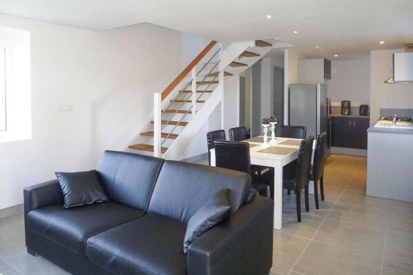 A modern living room with a black leather sofa, a dining table with chairs, and a kitchen area with a refrigerator and stove.