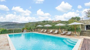 A swimming pool with lounge chairs and umbrellas, surrounded by stone walls and greenery, with a view of hills in the background.