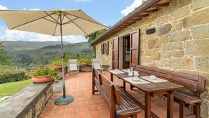 A patio with a table and chairs, a large umbrella, and a stone building with a view of the hills.