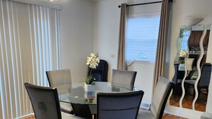 Un coin repas avec une table en verre, quatre chaises, un lustre, une orchidée en pot et une vue sur la cuisine grâce à un miroir.