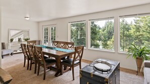 Un comedor con una mesa y sillas de madera, amplios ventanales con vistas a árboles, y una planta en maceta.
