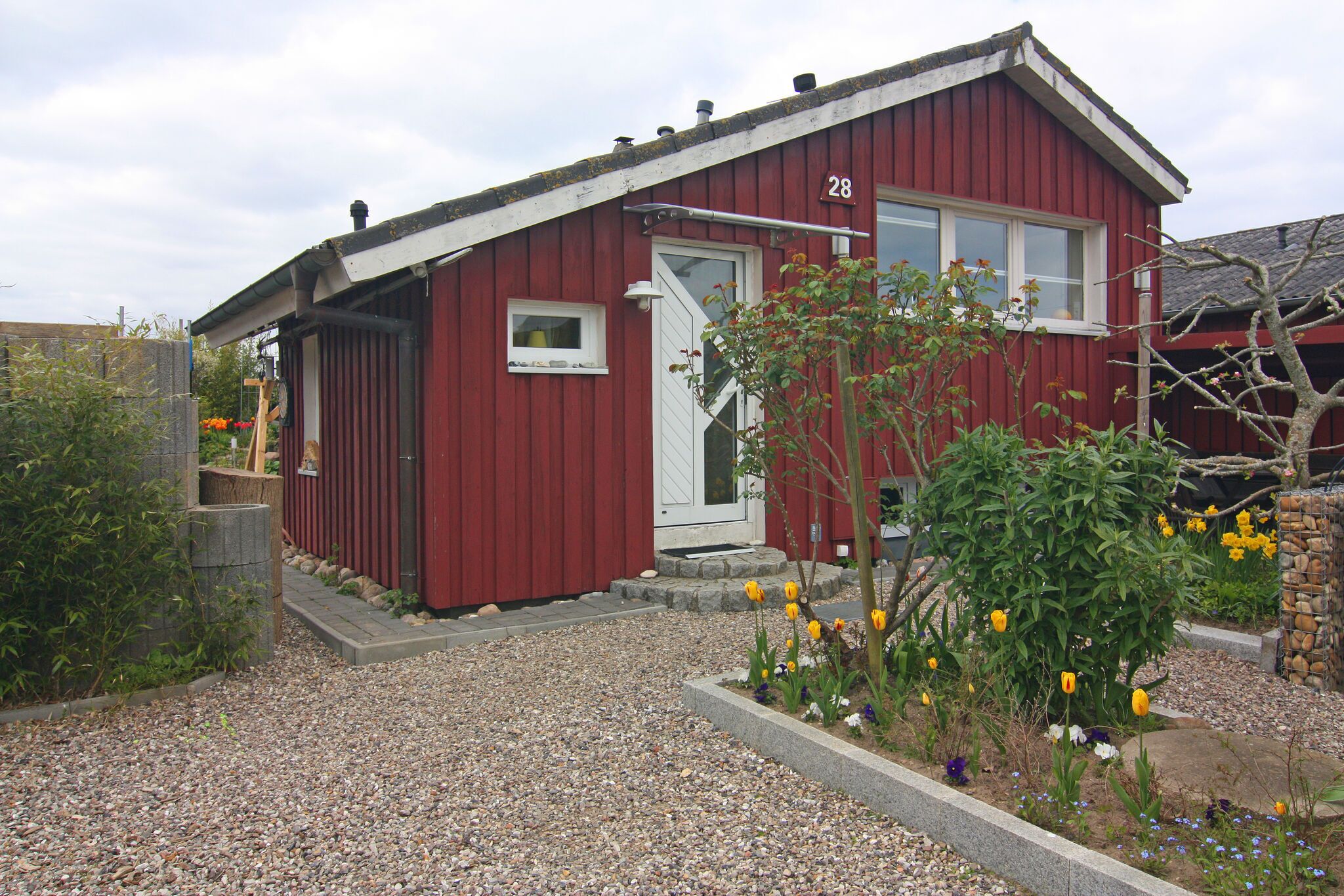 A red building with a white door and window, number 28, surrounded by gravel and a garden with flowers.