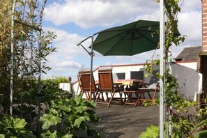 An outdoor patio with a table and chairs, a green umbrella, and a garden with bamboo and leafy plants.