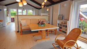 Una sala de estar acogedora con una mesa de comedor de madera, un sofá y un sillón mecedora. Hay una estantería y una ventana con vista a la vegetación.