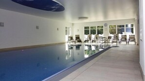 An indoor swimming pool with clear glass walls, a ceiling with a sky-like design, and a seating area with chairs and tables. - Appartements im Avalon Hotel Bellevue, Fehmarn (Fehmarn)