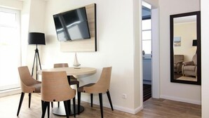 A dining area with a round table, four chairs, a flat-screen TV mounted on the wall, and a large mirror. - Appartements im Avalon Hotel Bellevue, Fehmarn (Fehmarn)