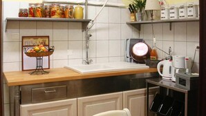 A kitchen with a wooden countertop, a sink, a kettle, and a coffee maker. There are jars of food on a shelf above the sink and a basket of fruit on the countertop.