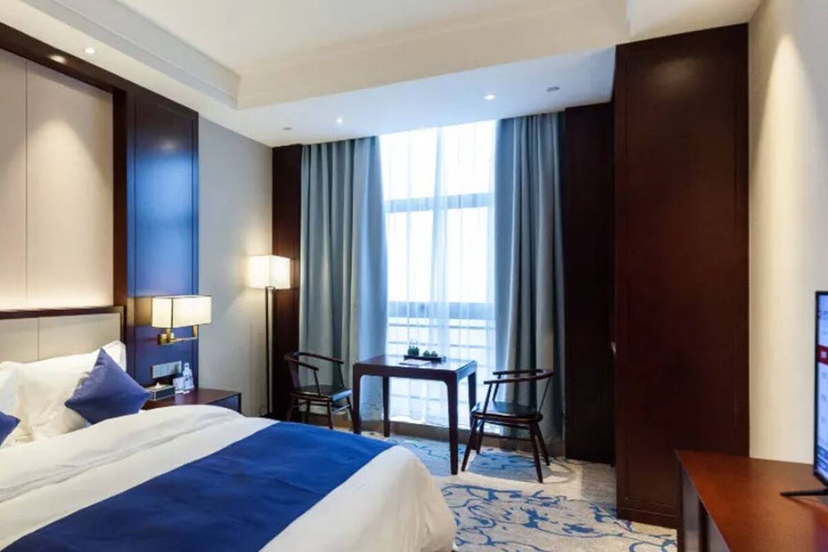 Ein Hotelzimmer mit einem großen Bett, einem Schreibtisch, zwei Stühlen, einer Lampe und Blick auf die Stadt durch große Fenster.