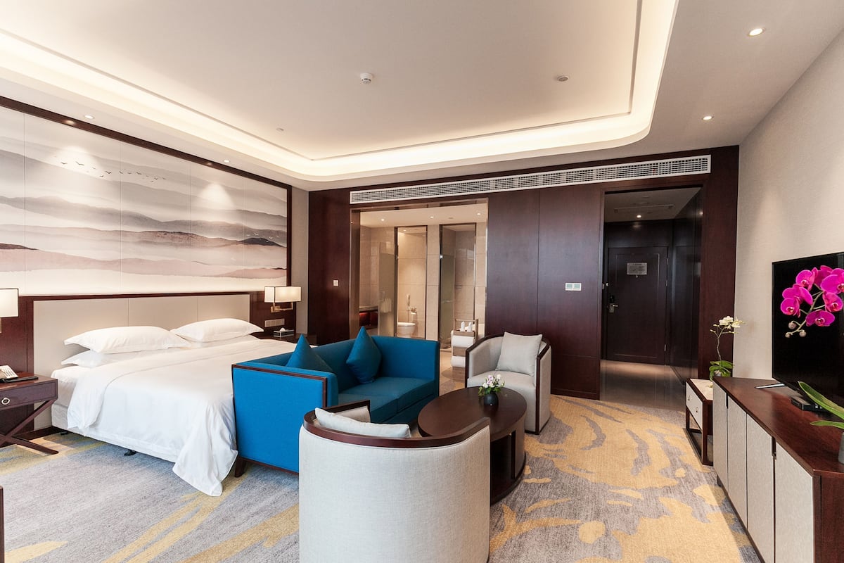 Ein modernes Hotelzimmer mit einem großen Bett, einem blauen Sofa, einem grauen Sessel, einem kleinen Tisch und einem Fernseher.