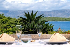 A table set with a glass of water, a glass of wine, and a folded napkin, with a view of the sea and mountains in the background.