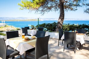Un espace extérieur aménagé pour les repas, avec des tables et des chaises, une vue sur la mer et une montagne en arrière-plan.